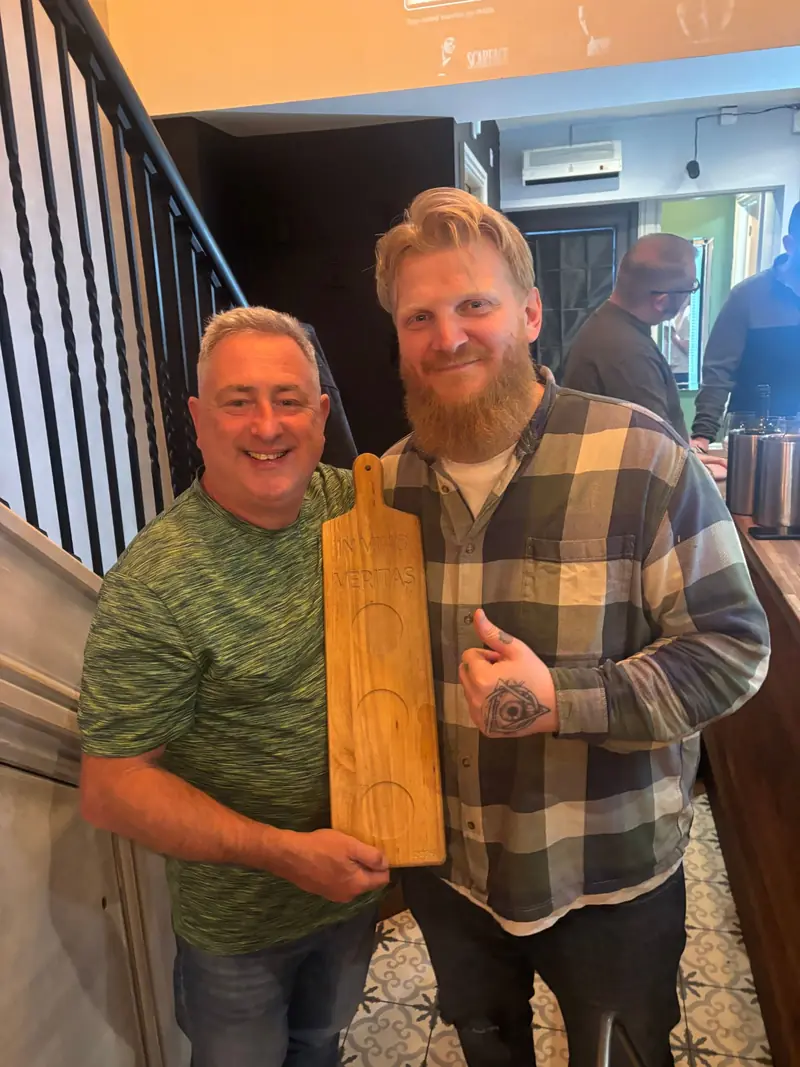 Phil and Stew holding a long wooden tapas board engraved with In Vino Veritas